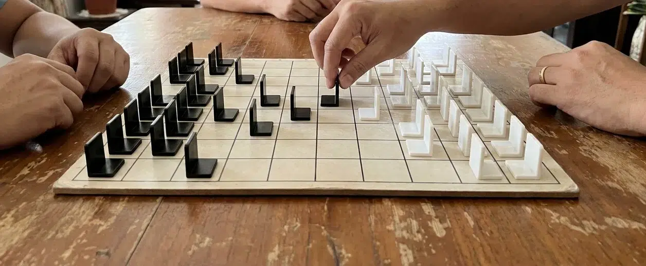 A game of the generals in progress on a wooden table: black and white upright pieces facing away from the opponent on a 9 by 8 grid board, and a player moving a piece one square